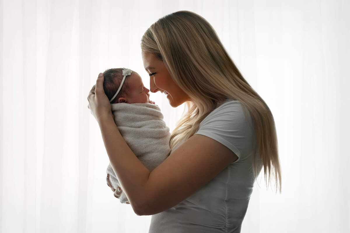 new mother holding her newborn baby girl  nose to nose wearing  white in front of a sheer white backlit curtain   at their VA Newborn  Photoshoot