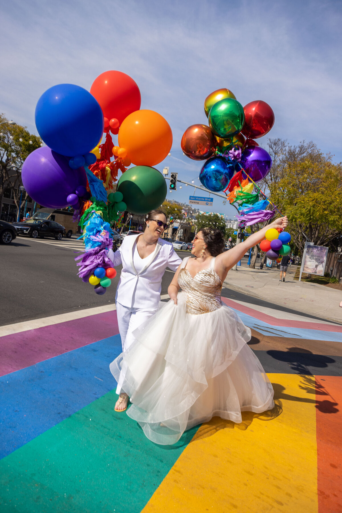 Two brides holding balloons and walking across a sidewalk painted with a pride flag.