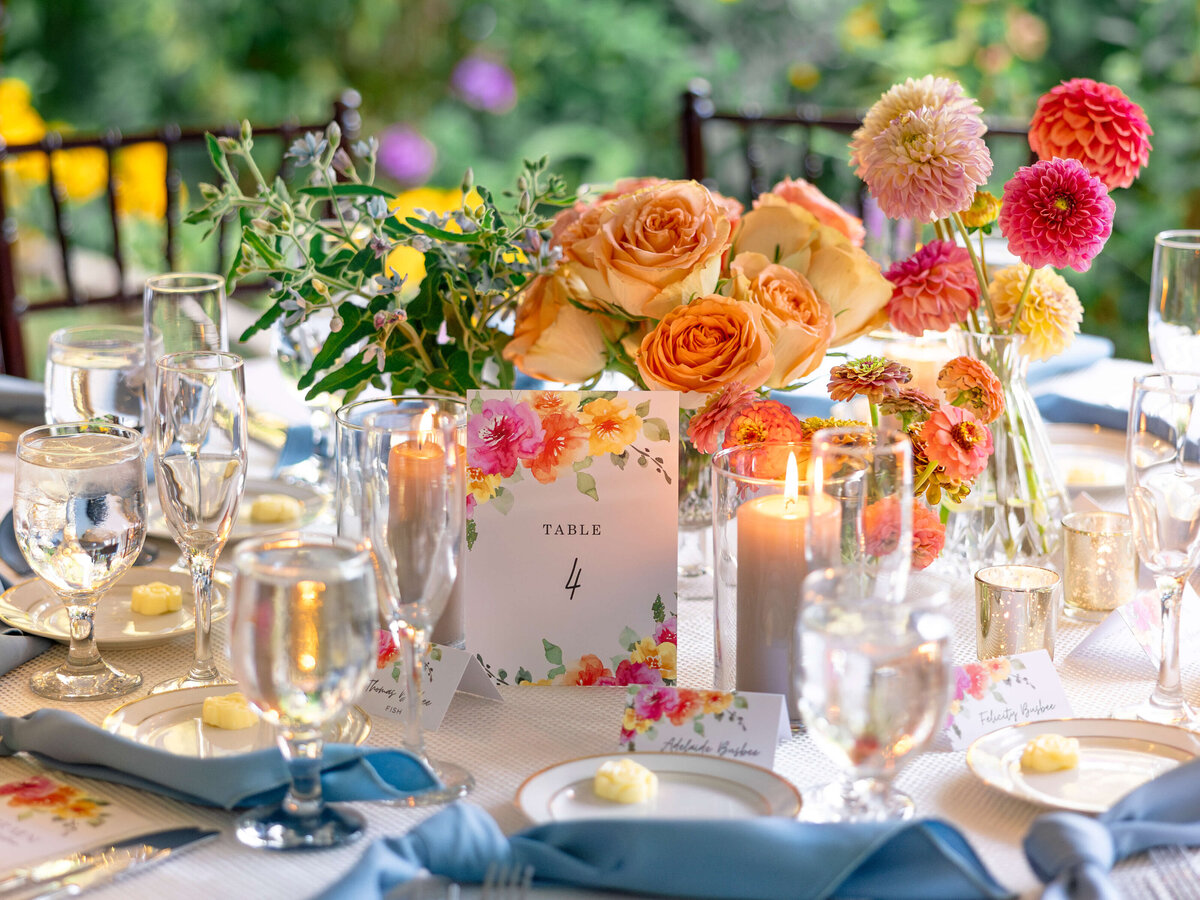 A colorful, elegantly set wedding table with a floral centerpiece, table number 4, candles, and glasses. The table is adorned with a white tablecloth and blue napkins, surrounded by nature.
