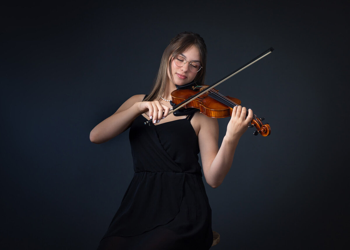 studio portrtait of a teenage girl in a black dress playing her violin with her eyes closed
