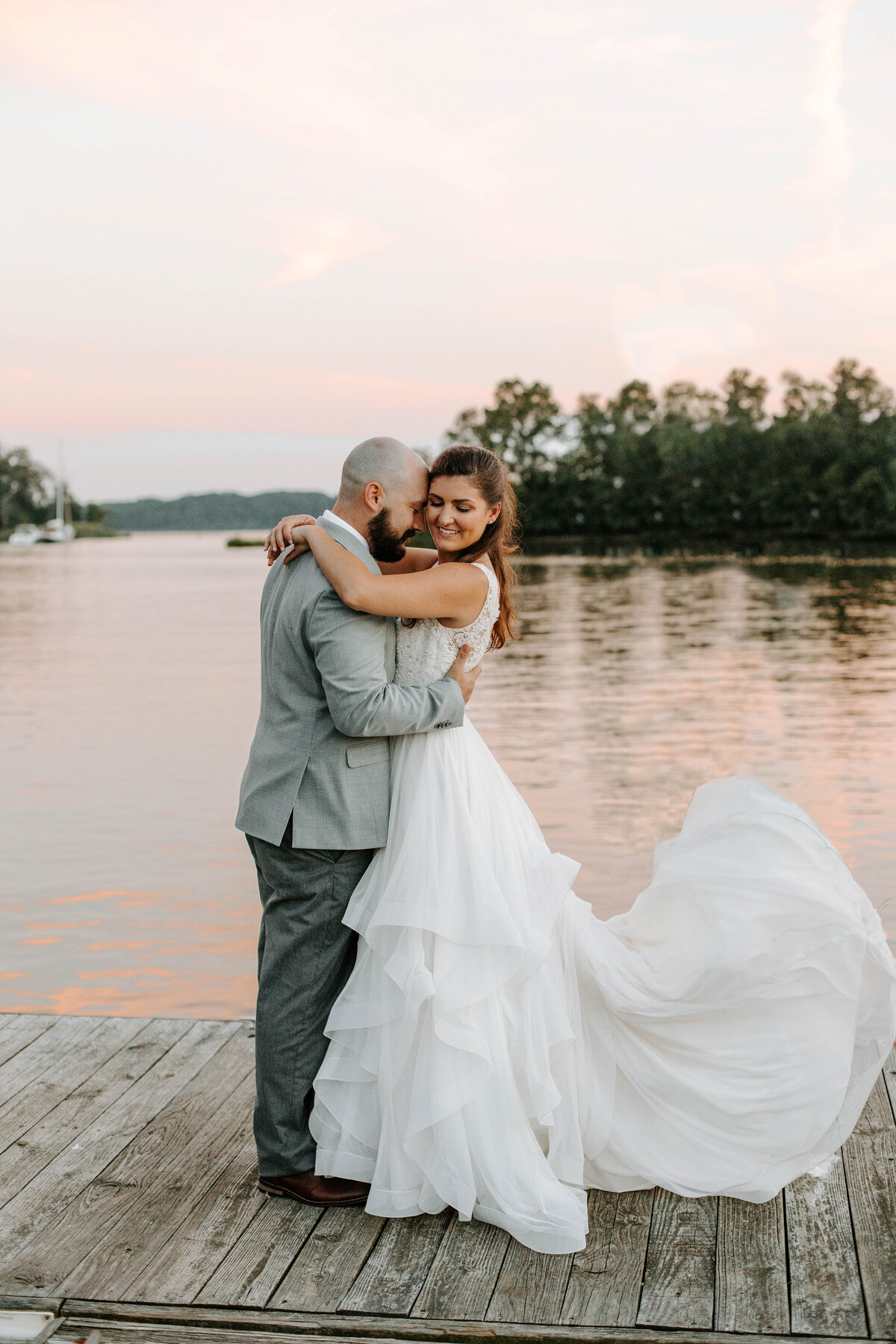A bride and groom stand on a pier in front of water that is reflecting a colorful sunset. The groom has his face towards the bride as the bride looks at the camera while her dress flows in the wind