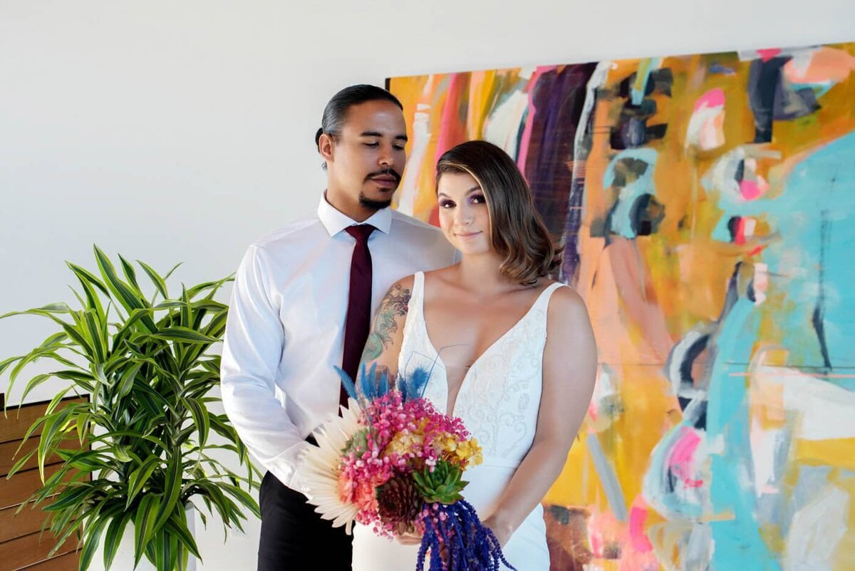 a bride and groom holding a very colorful bouquet stand in front of a vibrant mural by therese murdza