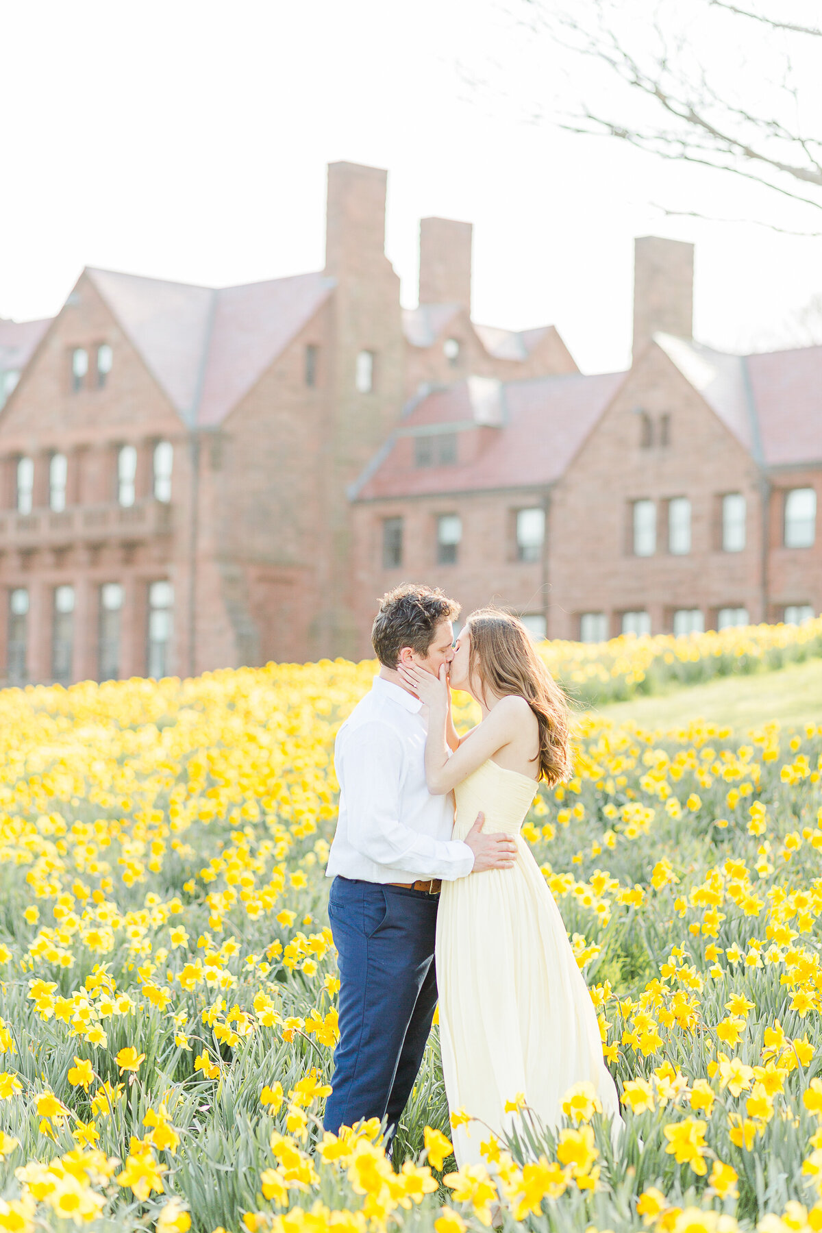 Man and woman stand amongst the daffodils at Salve Regina Cliff Walk. They are facing each other with their arms wrapped in an embrace and sharing a kiss. Salve Regina stone buildings are featured prominently in the background. Captured by best RI wedding photographer Lia Rose Weddings