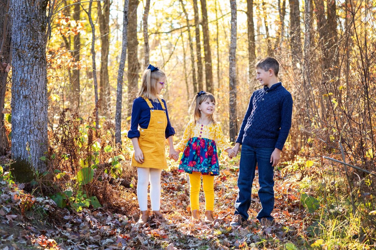 Three young siblings holding hands interacting with one another outdoors in the fall