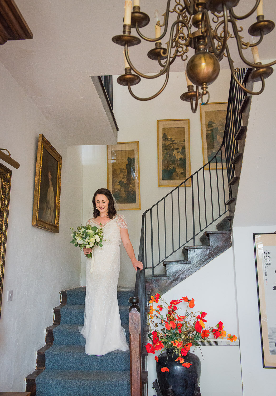 bride wearing a vintage, beaded wedding dress with sleeves walking down the stairs with blue carpet while holding a white flower bouquet