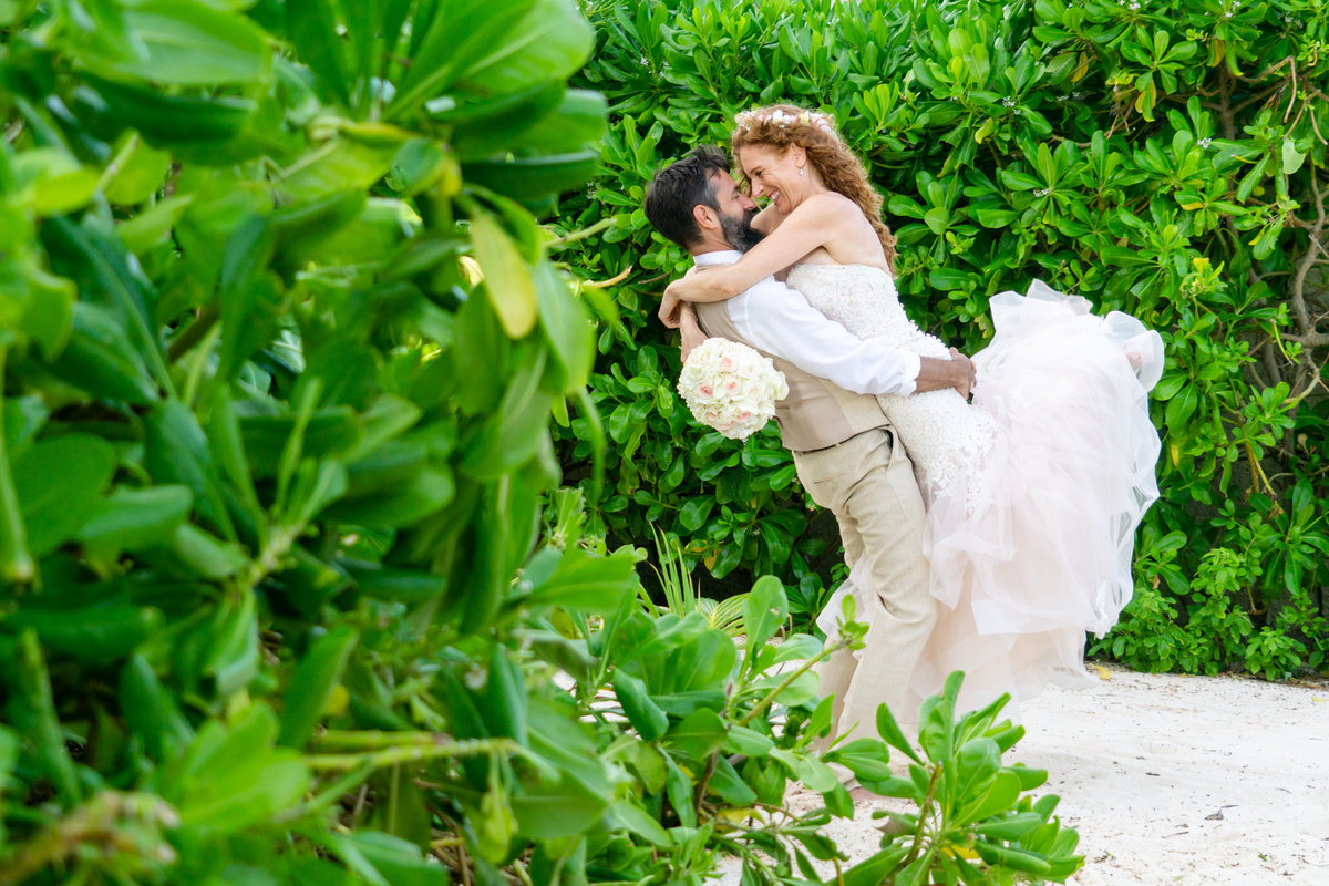 Groom holding wife with her feet kicked back in front of green bushes in Playa del Carmen