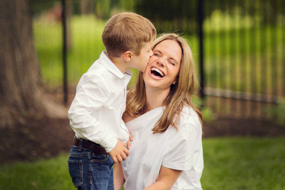 Back yard session and mom is laughing at the camera as he son plants a big kiss on her cheek.