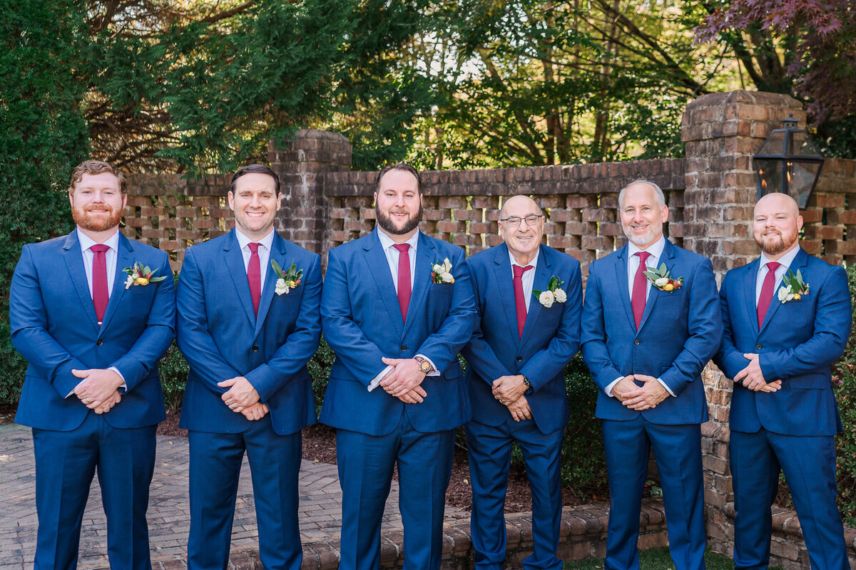 A groom and his six groomsmen in blue tuxedos under the fall leaves enjoying their Raleigh wedding photography session with JoLynn Photography 