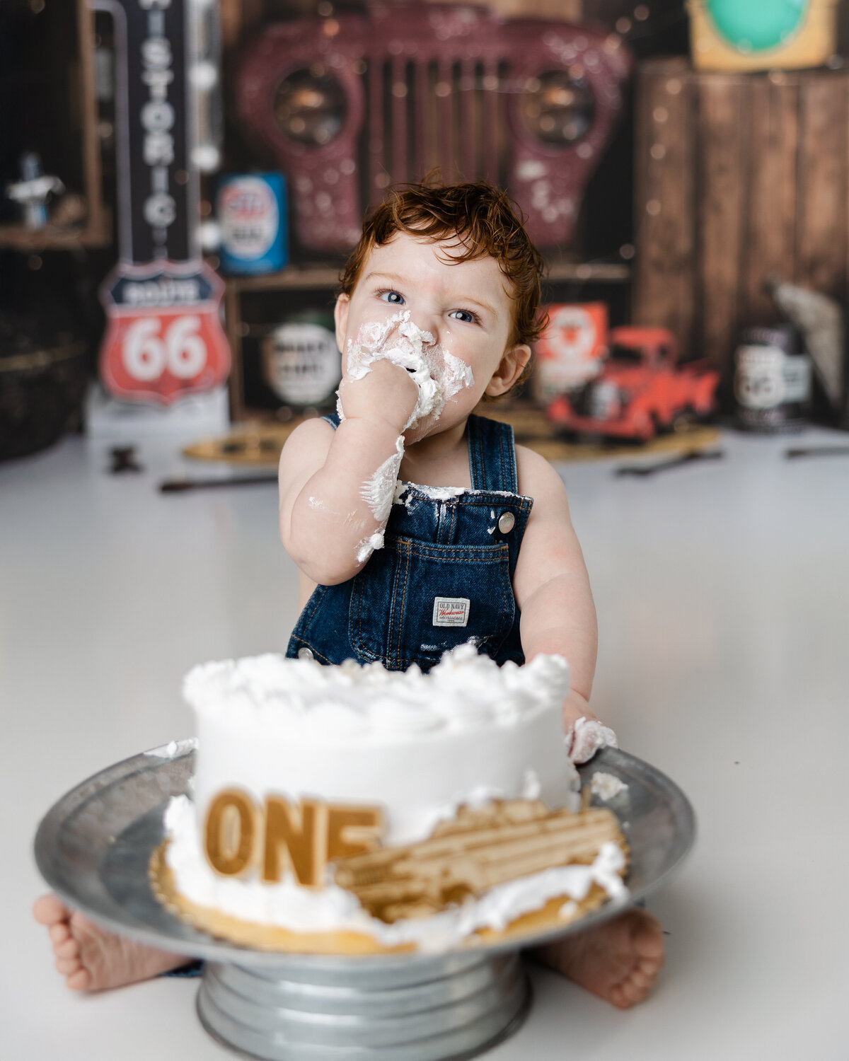Smash cake moment with a one-year-old boy enthusiastically tasting his cake in front of Route 66-inspired decorations. Photograph by Yucaipa cake smash photographer Kaitlyn Dawn Photography.