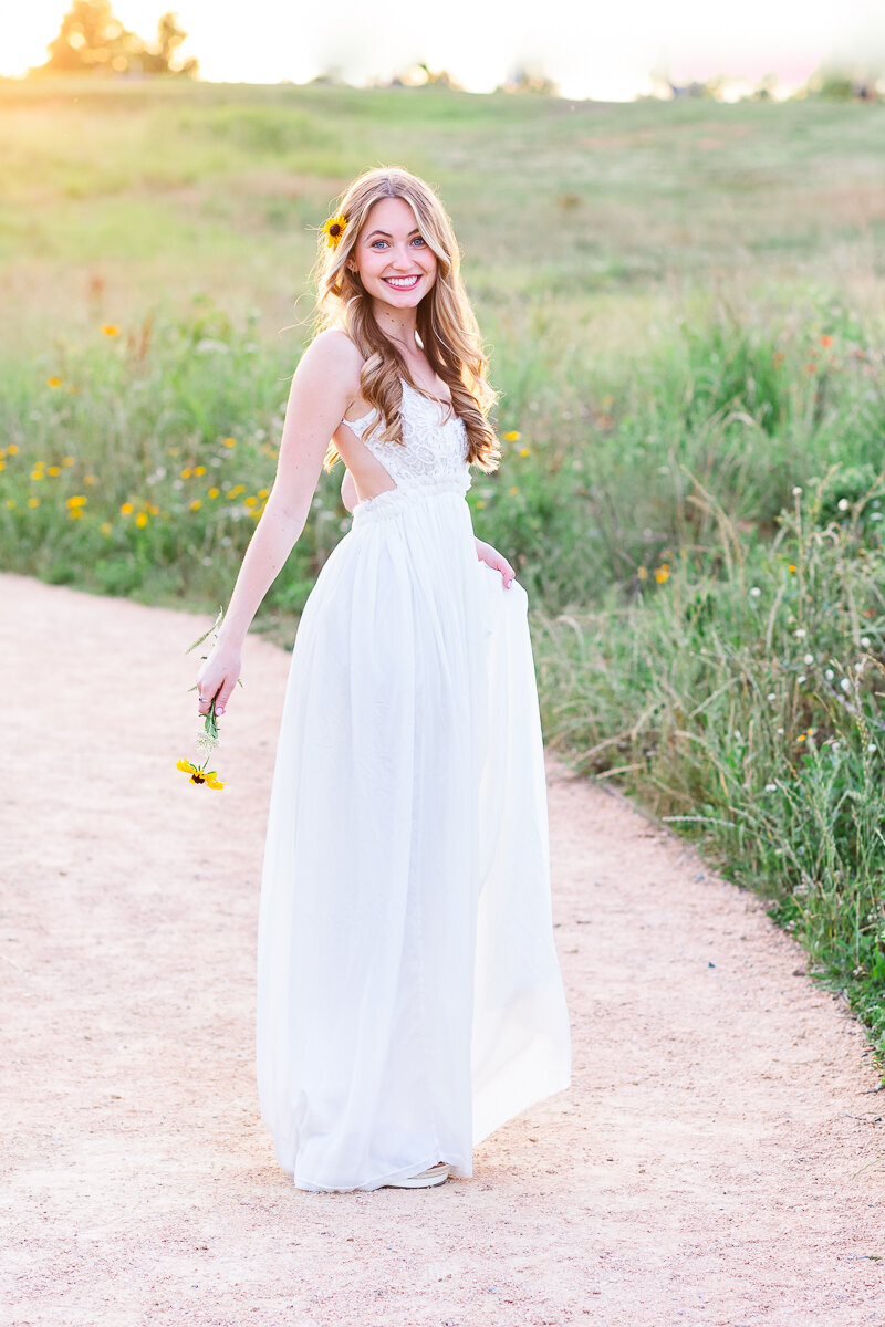 Senior girl with long white dress walking down the field during golden hour photo shoot.