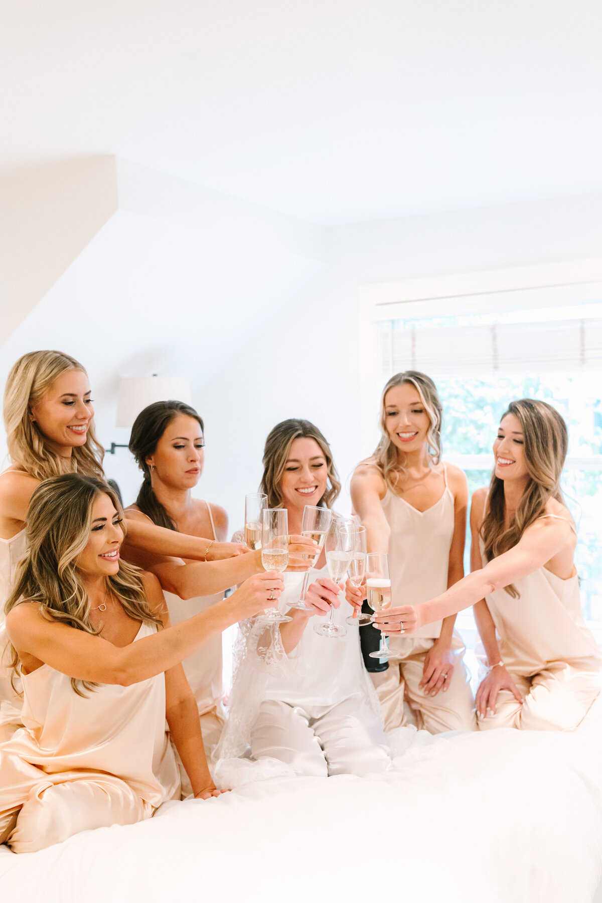 bride with bridesmaids cheersing champagne while sitting on a bed in soft silk pajamas.