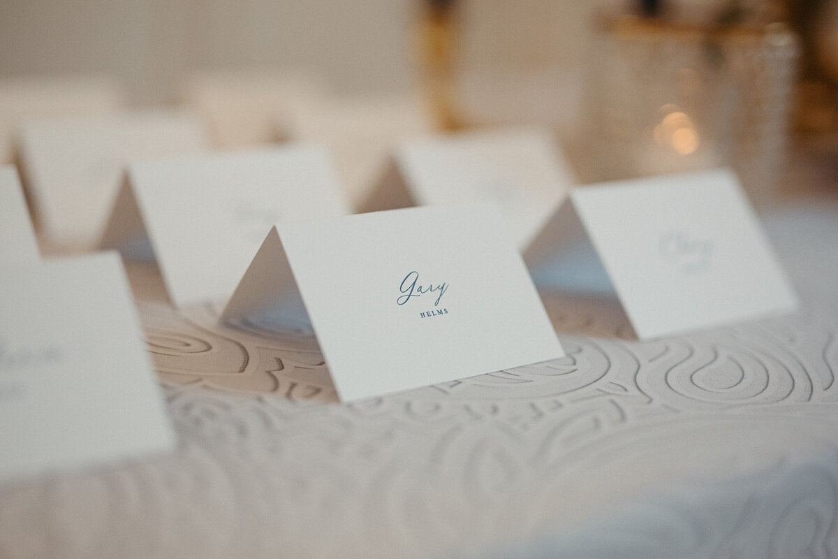 White place cards with cursive black font atop a table with white linen and a candle.