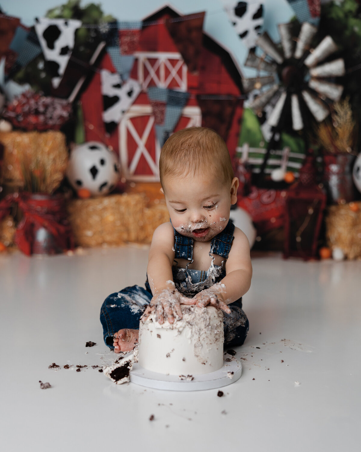 Baby boy in denim overalls smashing a white frosted cake during his farm-themed first birthday session, with a barn and hay backdrop.
Photograph by Yucaipa cake smash photographer Kaitlyn Dawn Photography.