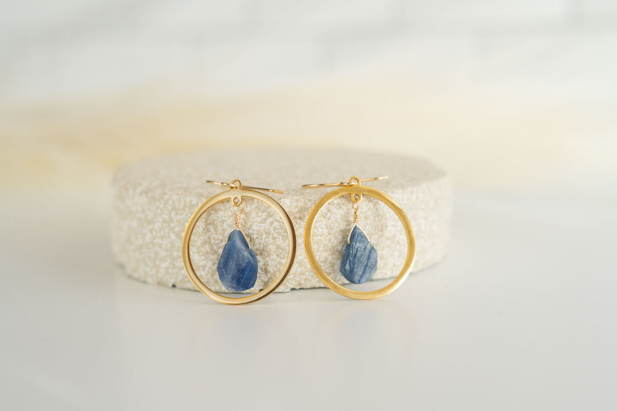 Gold hoop earrings with blue gemstone pendants displayed on a white textured surface.