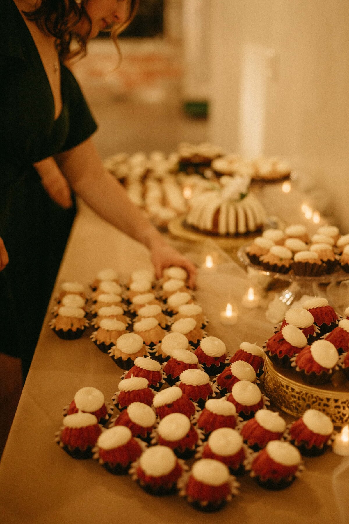 A wedding planner in a green dress arranges an assortment of cupcakes on a table lit by candles at an event.