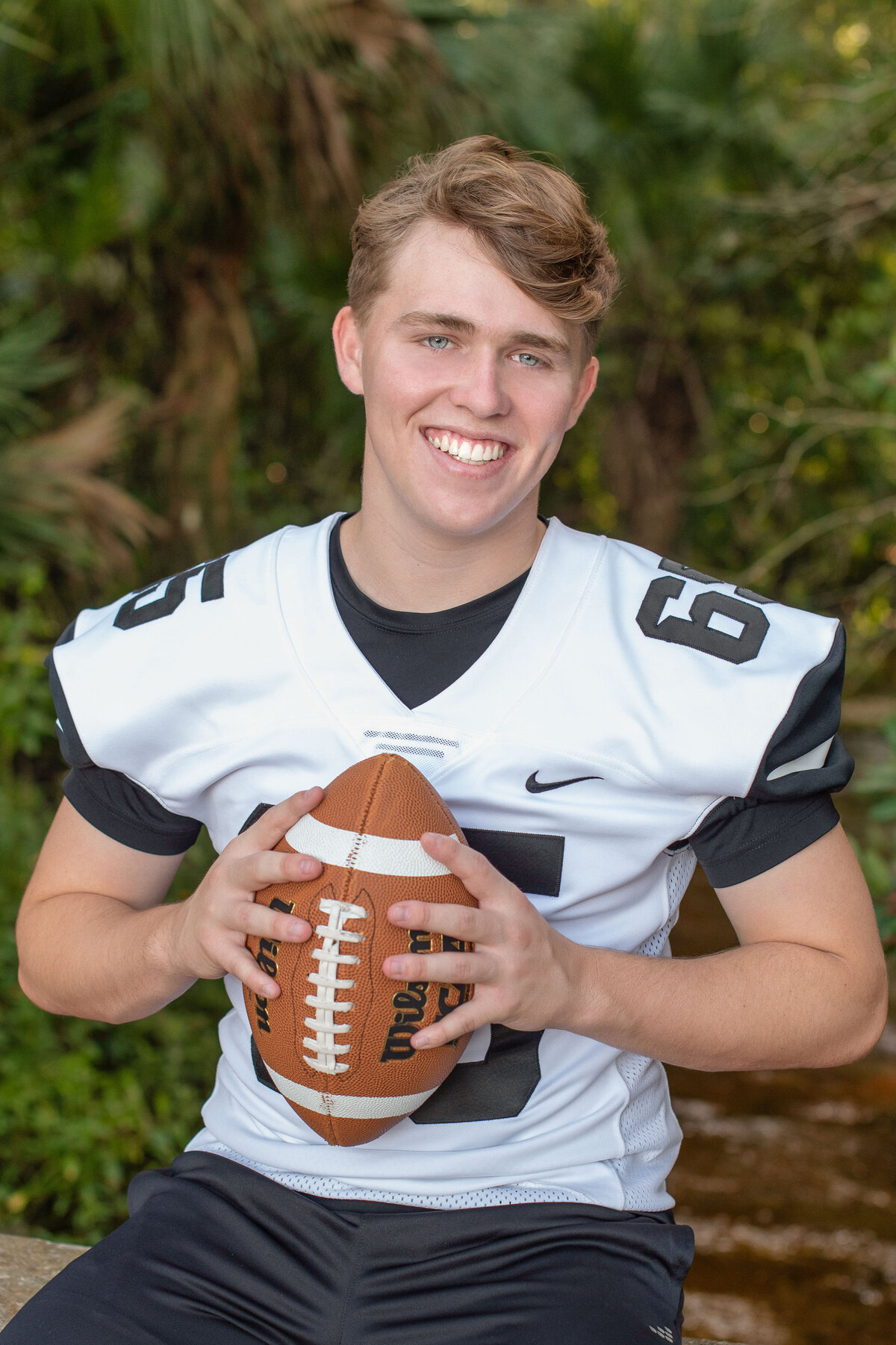 High school senior boy wearing a football jersey and hold a football sits on a bench and smiles at the camera.