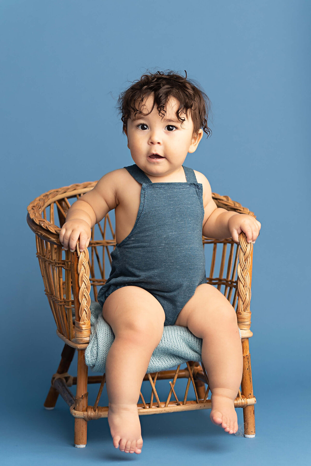 baby boy wearing blue overalls and in a chair looking serious with his adorable curly brown hair. Taken by portland photographer Ann Marshall