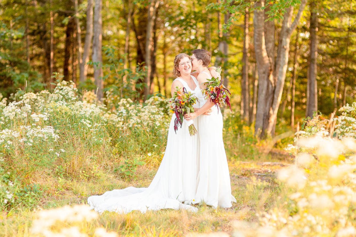 Lesbian brides laugh as they kiss in a forest.