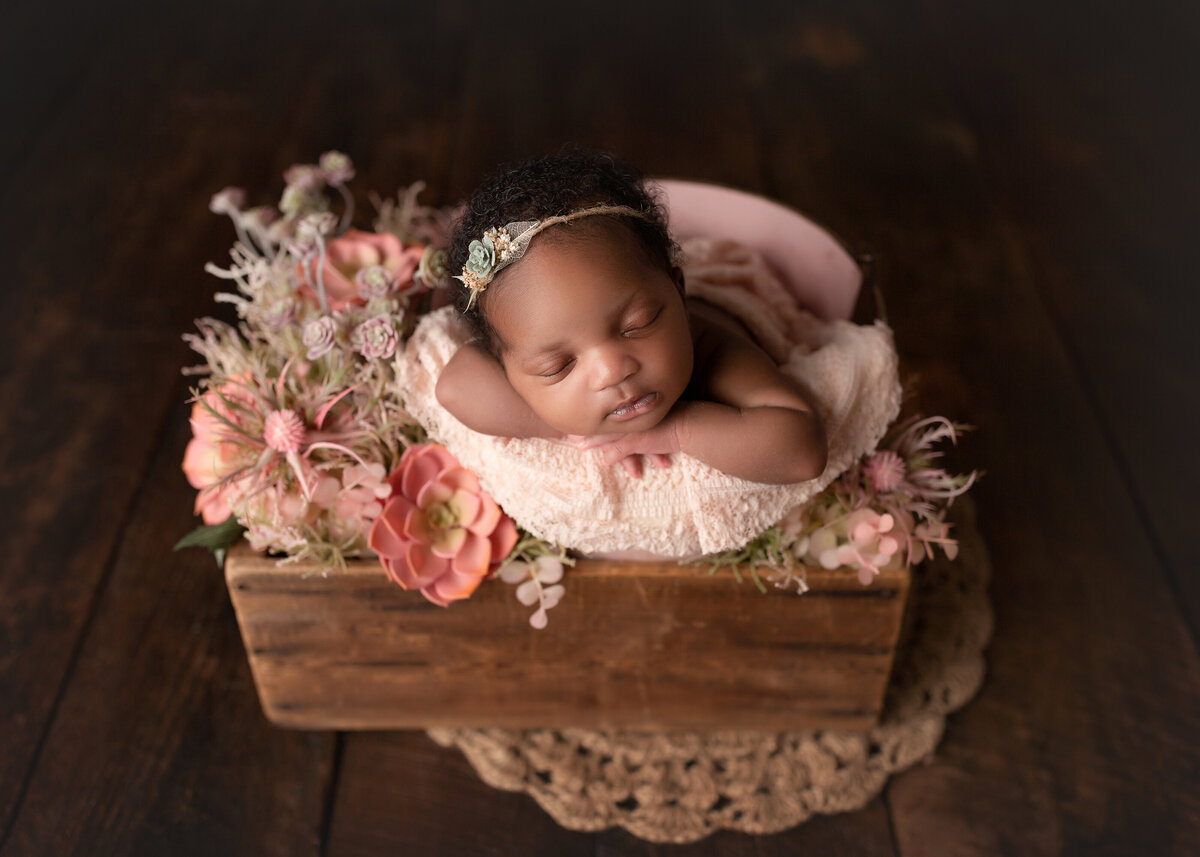 Boynton Beach newborn photoshoot - Black Baby is sleeping in a crate filled with blush and coral flowers. Baby's hands are folded under her chin and resting her cheek on her left arm. Baby is wearing a delicate floral headband.