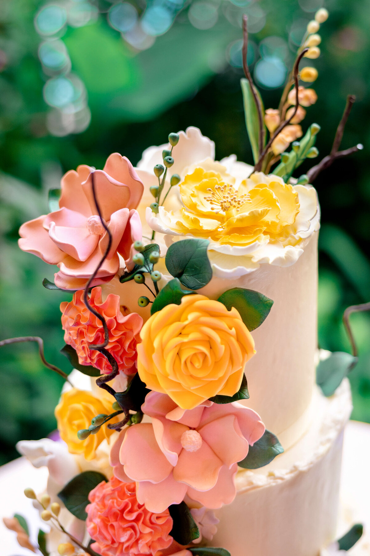 A beautifully decorated two-tier cake adorned with intricate, realistic sugar flowers in soft pastel shades of yellow, pink, and peach. The flowers cascade elegantly down the side against a lush, blurred green background.