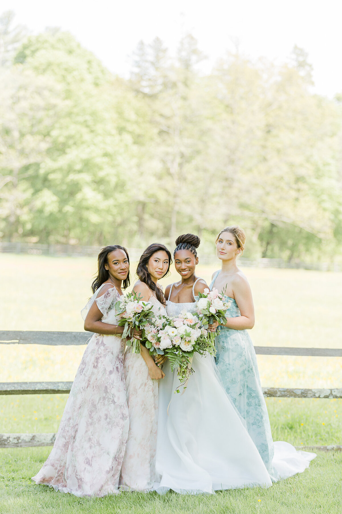 Bride is standing in a field with her three bridesmaids. All are holding their flowers in front of them. Captured by best Rhode Island Wedding Photographer Lia Rose Weddings.