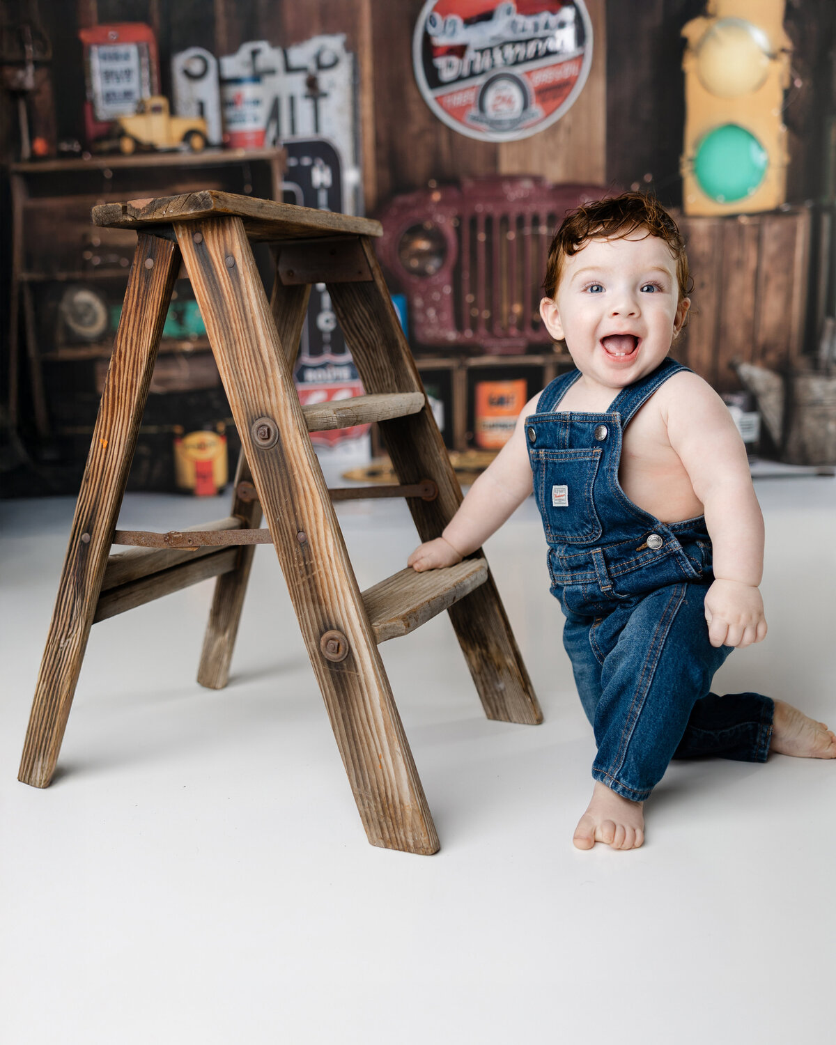 Smiling baby boy in denim overalls holding onto a wooden ladder during his truck-themed first birthday session.
Photograph by Yucaipa cake smash photographer Kaitlyn Dawn Photography.