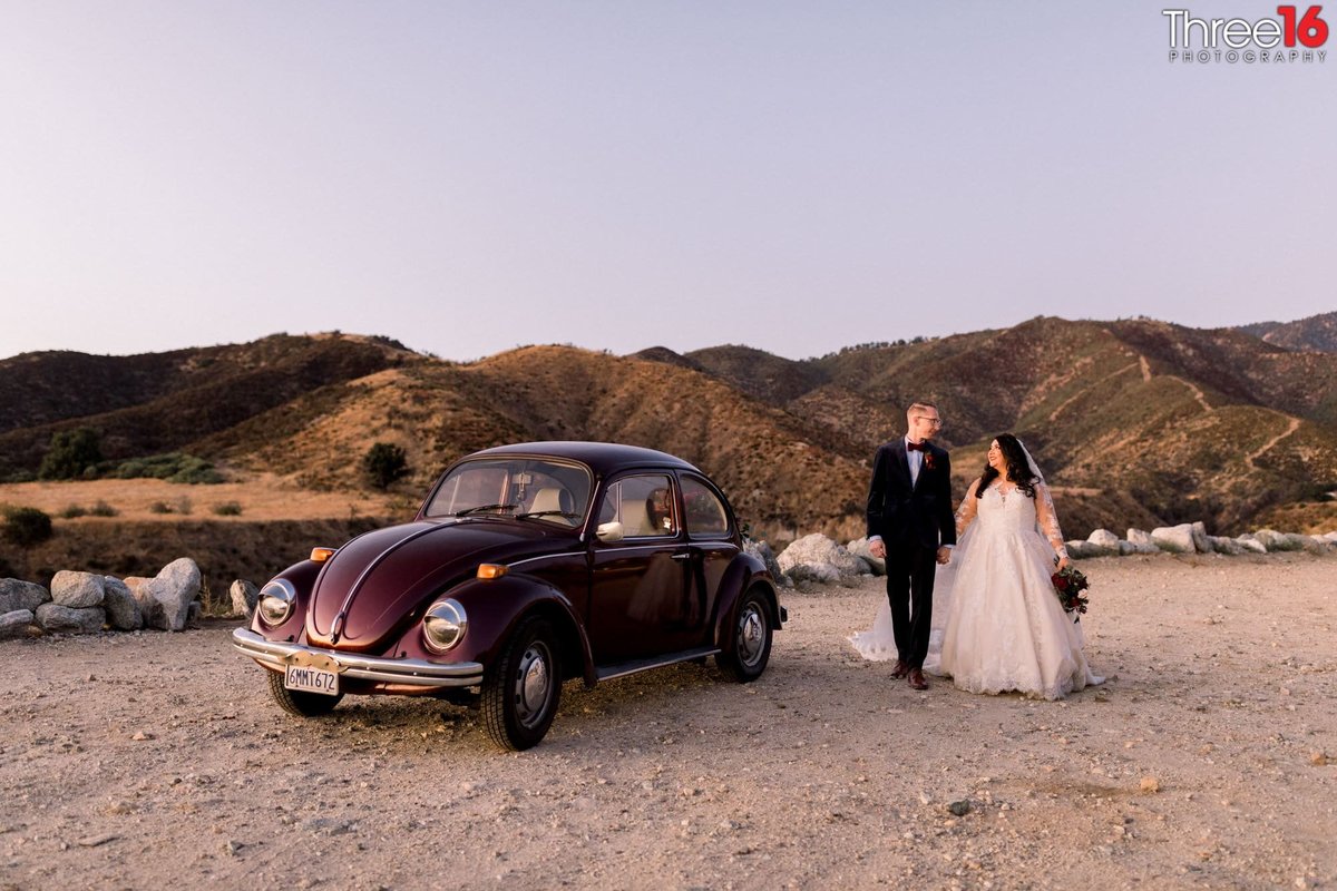 Bride and Groom walk holding hands on the dirt lot next to a maroon colored Volkswagon Bug car