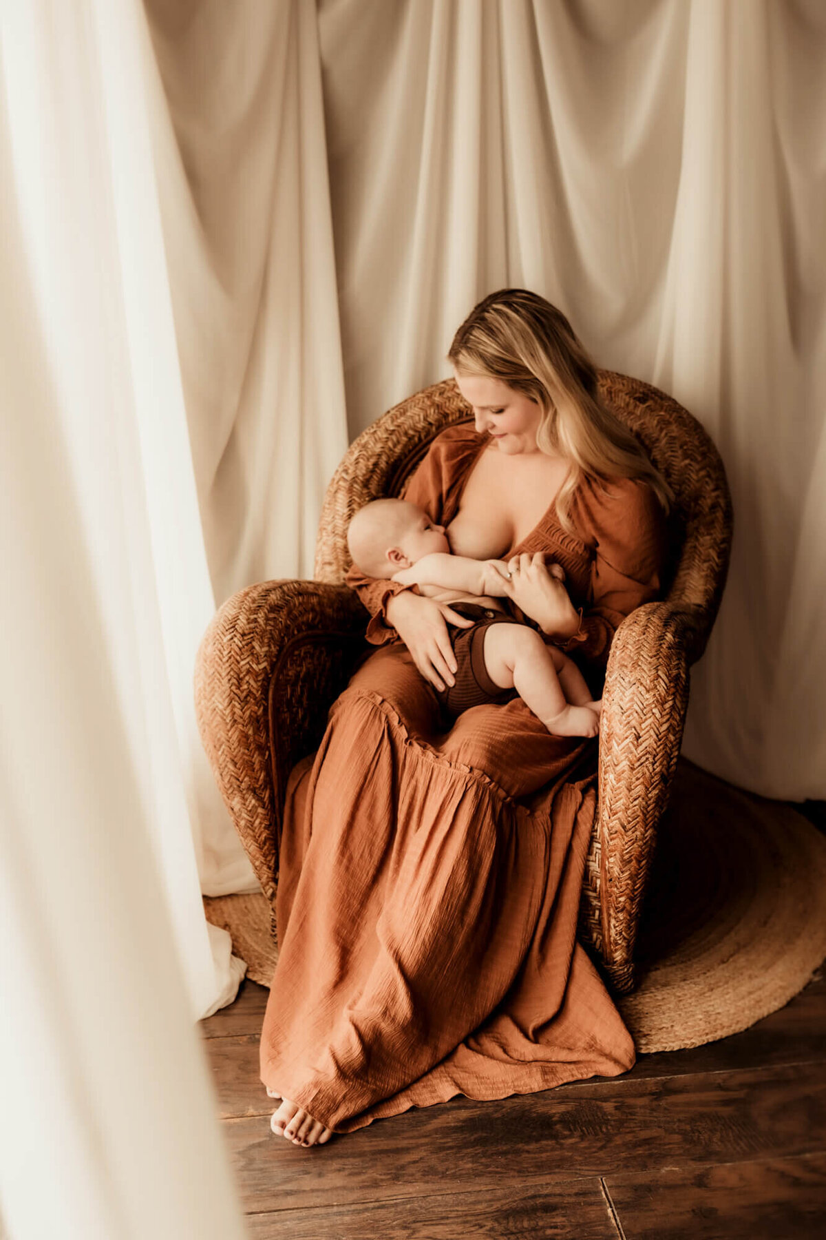 Mother wearing a rust colored dress breastfeeding her baby boy near a window.