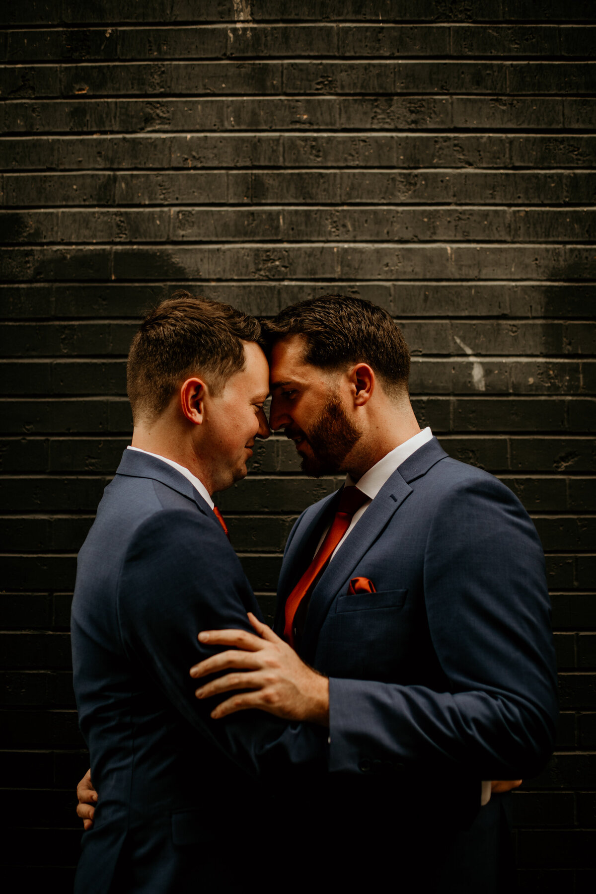 two grooms in blue suits hodling each other in front of a black brick wall in Albuquerque