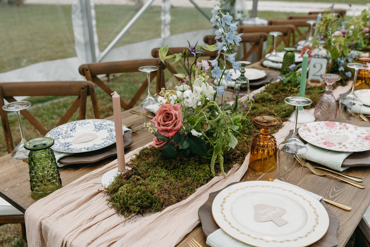 Garden-style wedding tablescape featuring a natural moss table runner, blush taper candle, and a romantic floral centerpiece with pink roses, blue delphinium, chamomile, and textured greenery. Mismatched vintage plates, amber glassware, and soft pastel details create a whimsical, meadow-inspired design for an outdoor tented wedding reception in Northwest Arkansas, created by a garden-style wedding florist.