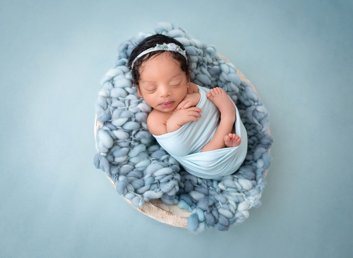 Baby girl wrapped in a blue swaddle with her hands and toes peeking out for a newborn photoshoot. Baby's hands are folded under her chin. She is wearing a light blue headband and sleeping atop of blue blankets.