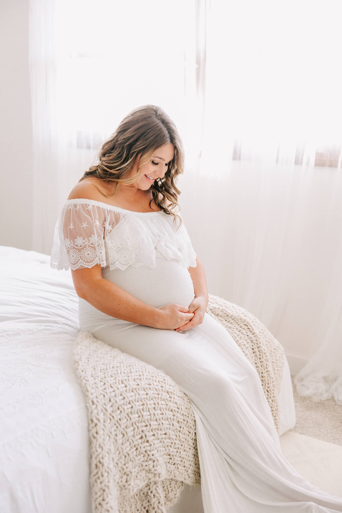 portland oregon woman sitting on bed in white dress smiling down at her pregnant belly