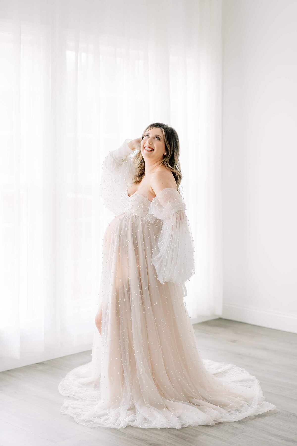 A brunette joyfully poses in  a tulle and pearl maternity gown as she looks up to the ceiling with her hand in her hair.