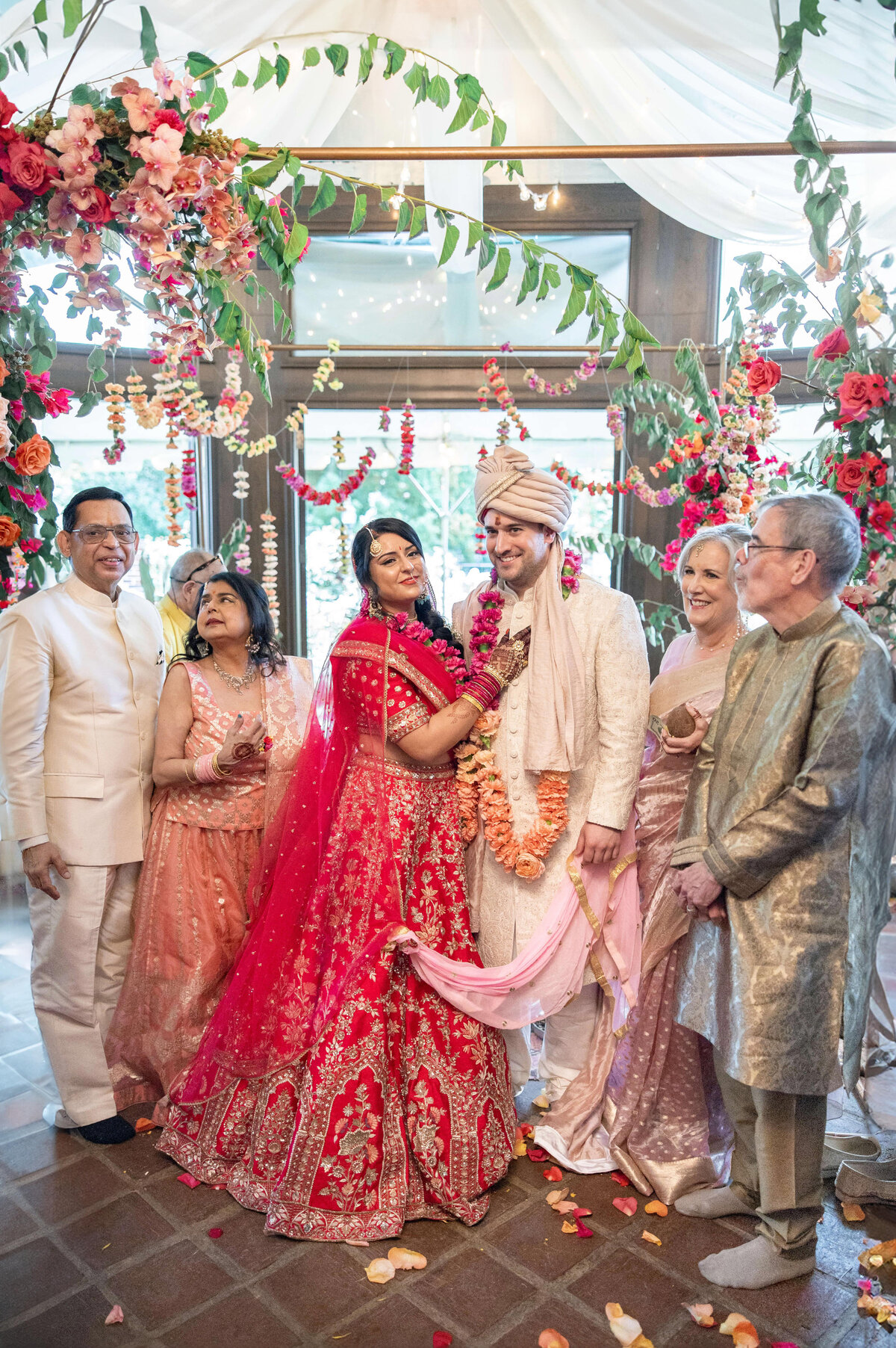 A joyful wedding scene with a couple in traditional attire surrounded by six smiling people. The bride wears a vibrant red lehenga and the groom is in a cream sherwani. The backdrop is decorated with colorful flowers and greenery.