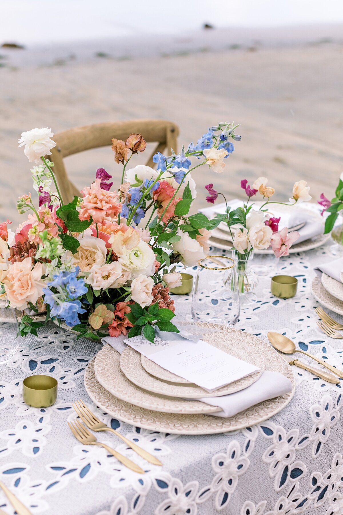 Gold, coral, blue, and blush tablescape with gold accents atop an eyelet tablecloth on beach
