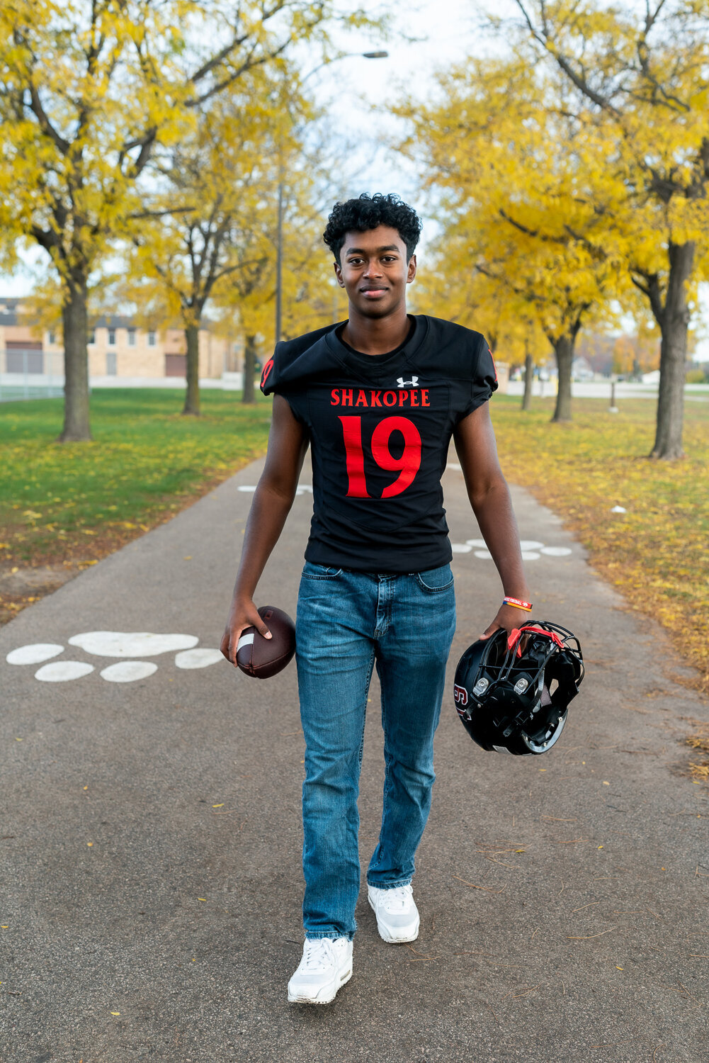 Senior boy walks down path in football jersey holding a football and helmet.