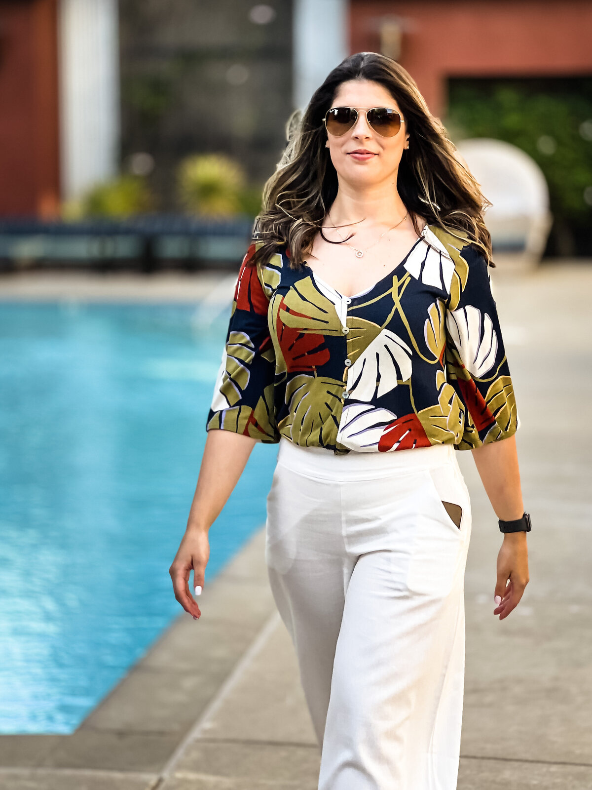 Woman walking close to a pool, wearing  a flare white pair of pants and a flower blouse