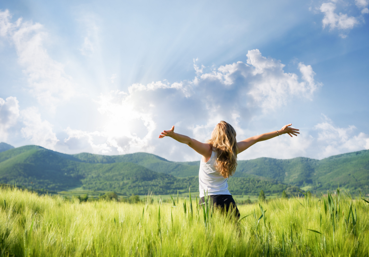 Woman stands in a field surrounded by mountains with her hands stretched to the sky.
