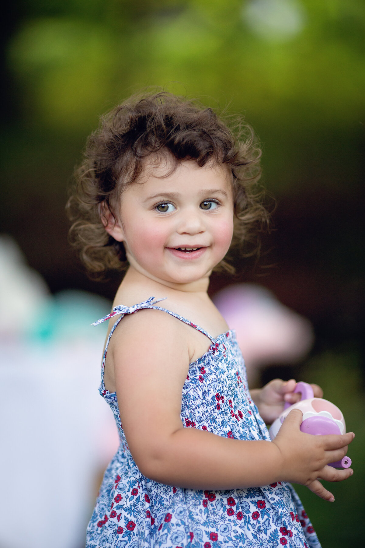 A toddler girl in a blue floral print dress plays with a toy in a park