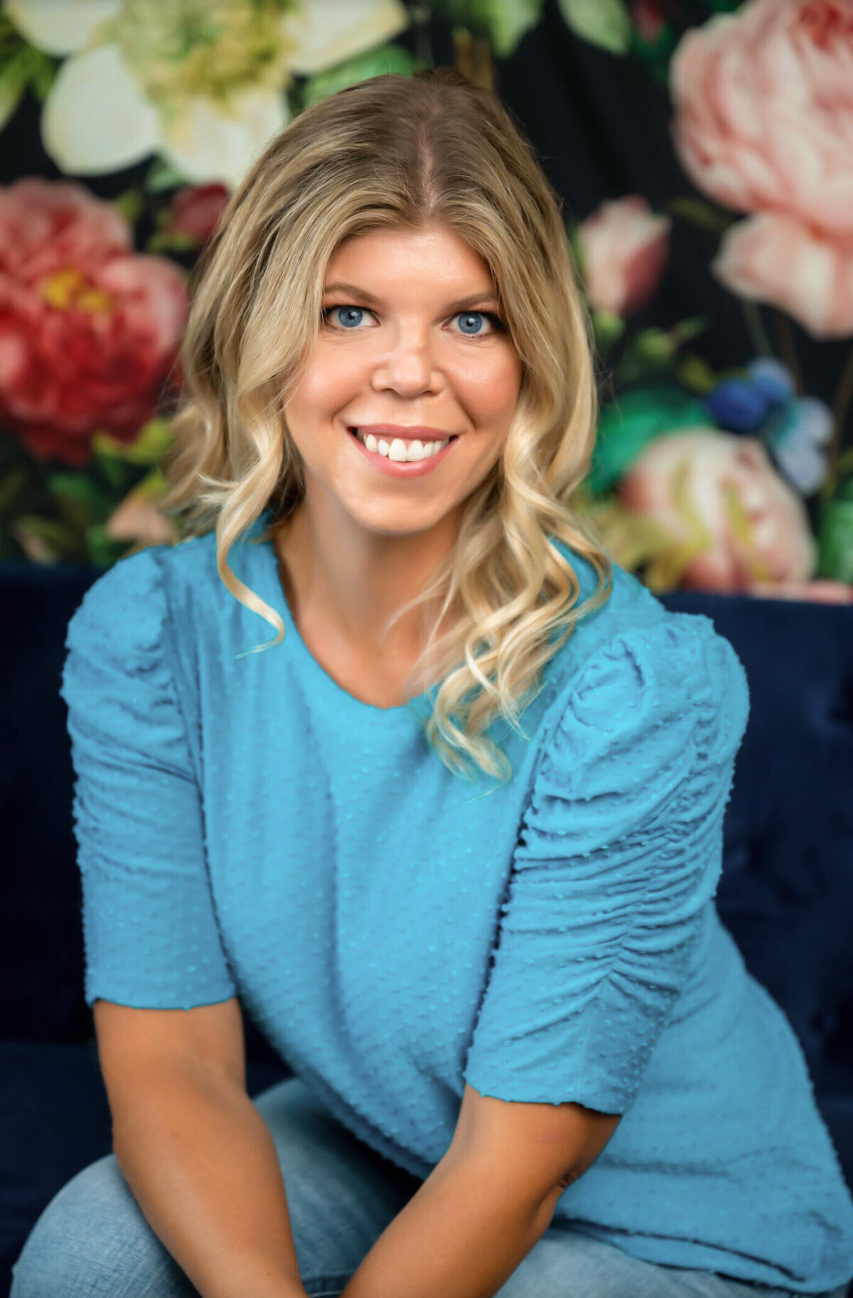 A woman with blonde hair and blue eyes wearing a blue shirt in front of a floral background posing for a headshot