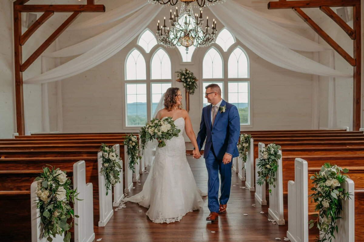photo of a bride and groom holding hands and walking down the aisle of a white chapel with chandeliers and wooden benches