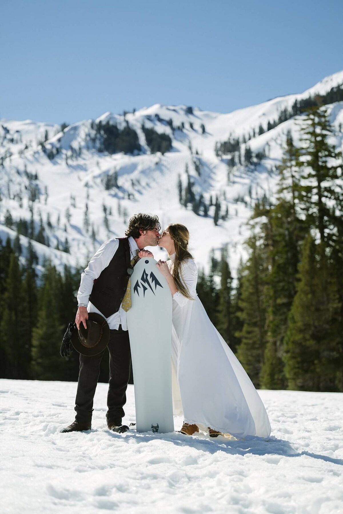 Couple Newlyweds kissing with a snowskate with snowy mountains in backdrop during winter elopement at Palisades Tahoe Alpine.