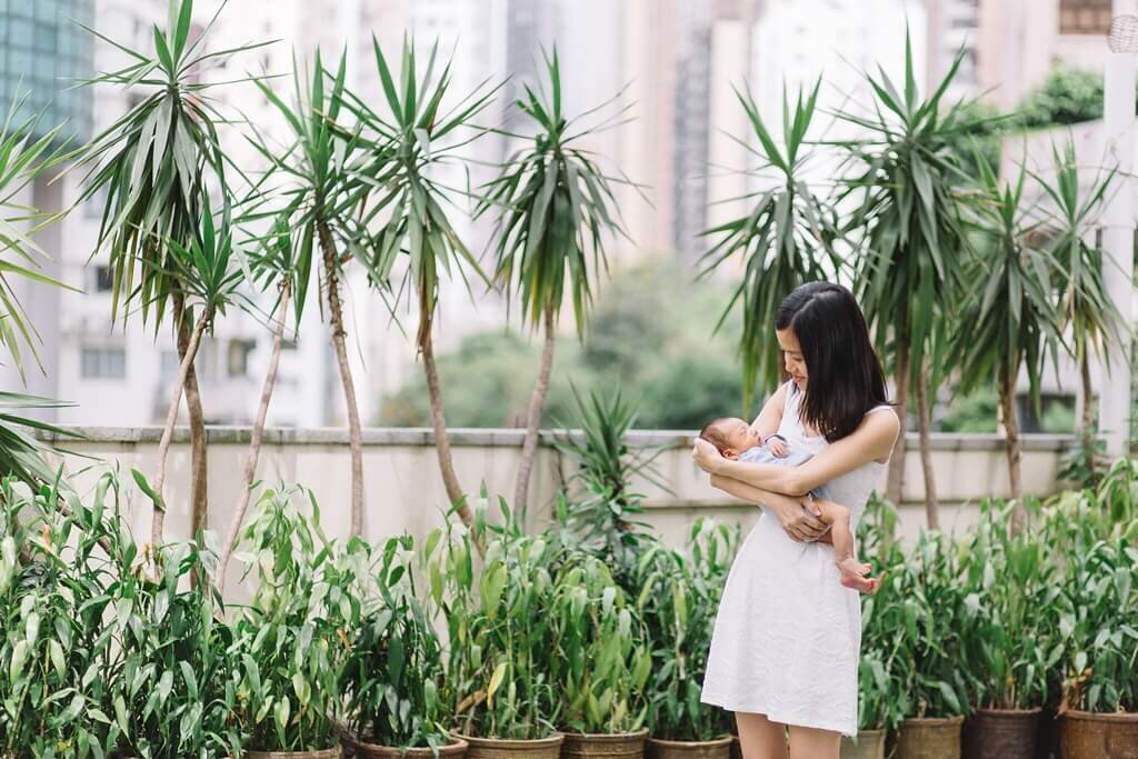 Mother outdoors holding her newborn with small potted palms trees lined up in the background.