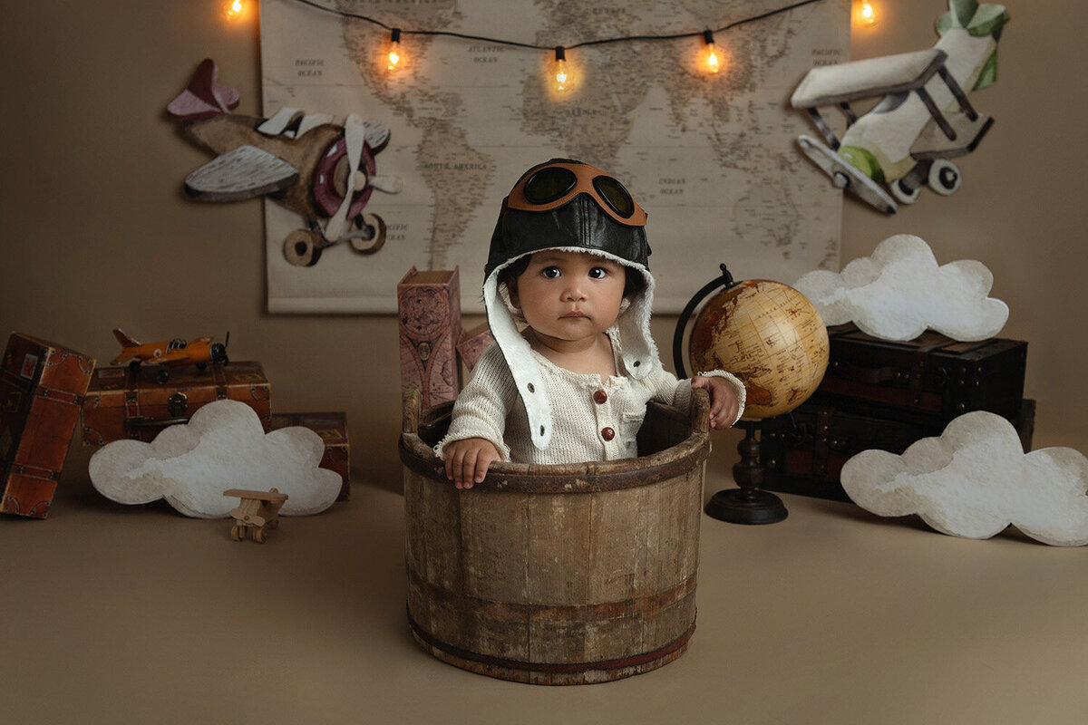 A young toddler dressed as a vintage aviator sits in a wooden bucket in a decorated studio