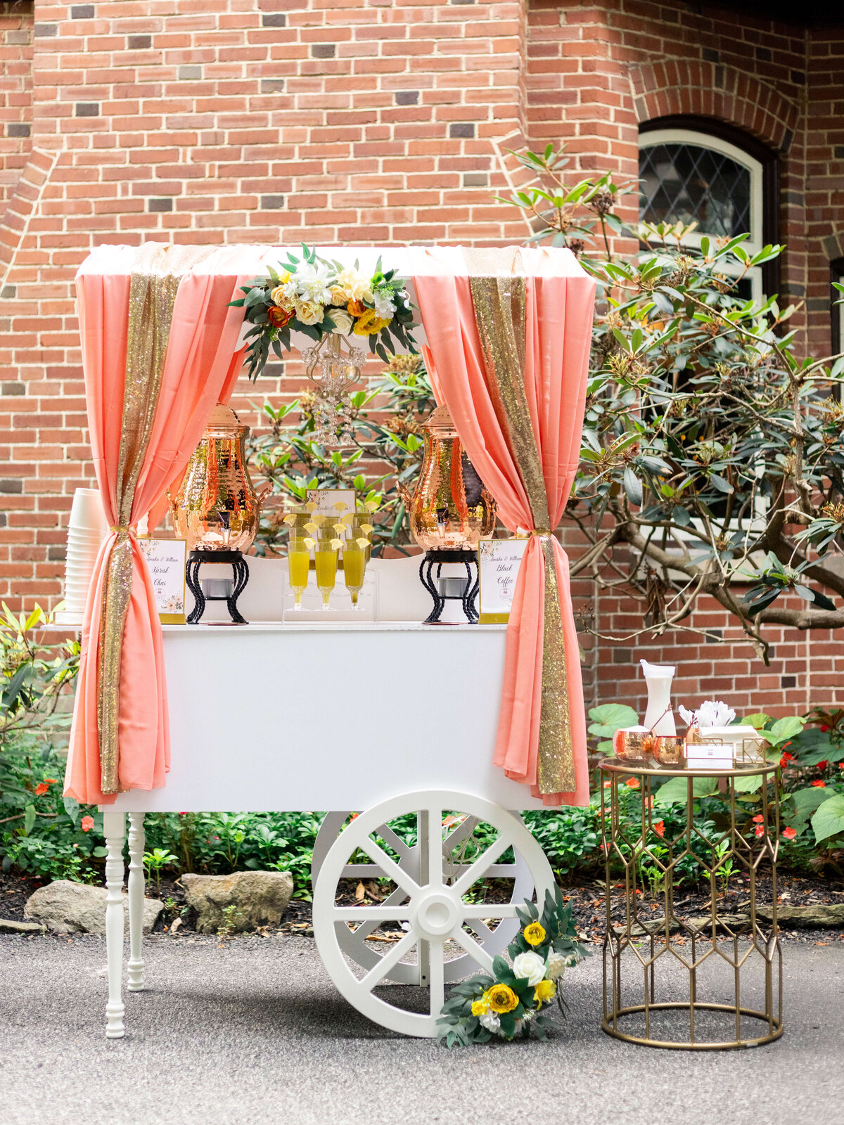 A charming white cart with large wheels draped in pink and gold curtains, adorned with floral arrangements and glassware, set against a brick building and greenery. A small side table holds additional decorative items.