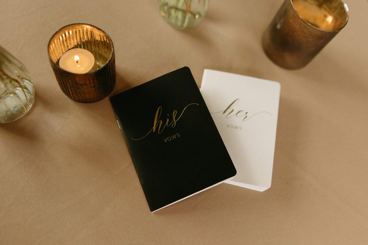 Two wedding vow booklets labeled "his" and "hers" on a table, surrounded by candle holders with lit candles, prepared by a wedding coordinator in Iowa.