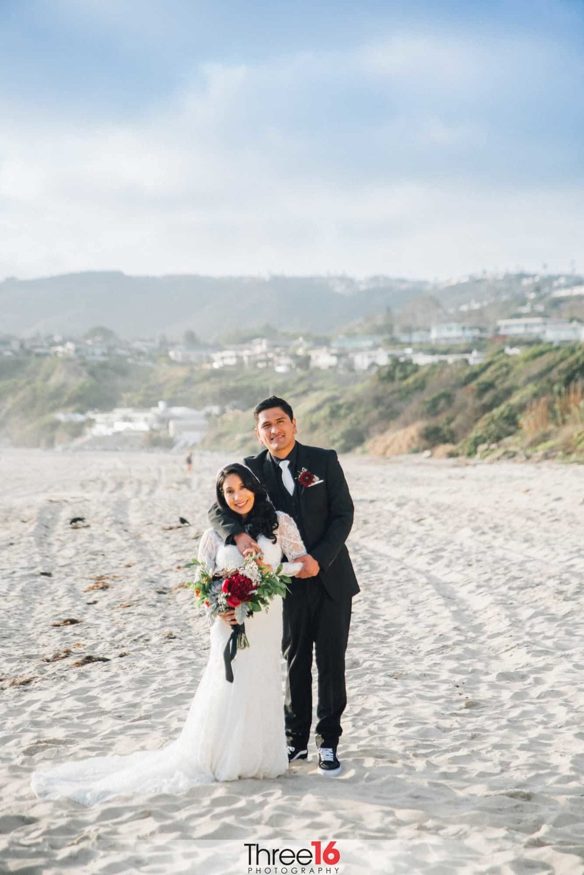 Groom embraces his Bride from behind while standing on the sands of Salt Creek Beach in Dana Point