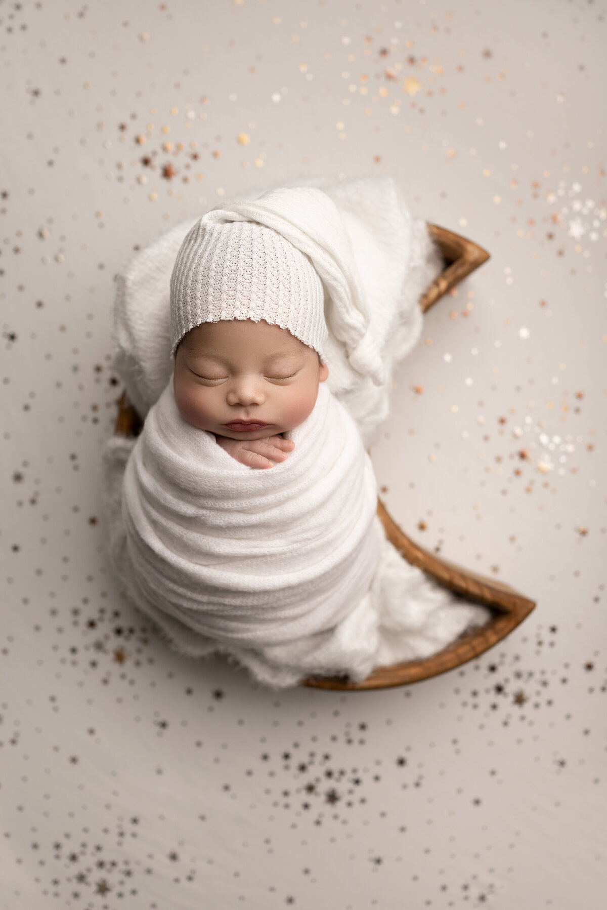 New Jersey's best newborn photographer, Katie Marshall, captures an aerial image of a sleeping newborn baby. Baby is swaddled in white with his fingers peeking out. He is wearing a long cap and laying in a moon-shaped bowl.
