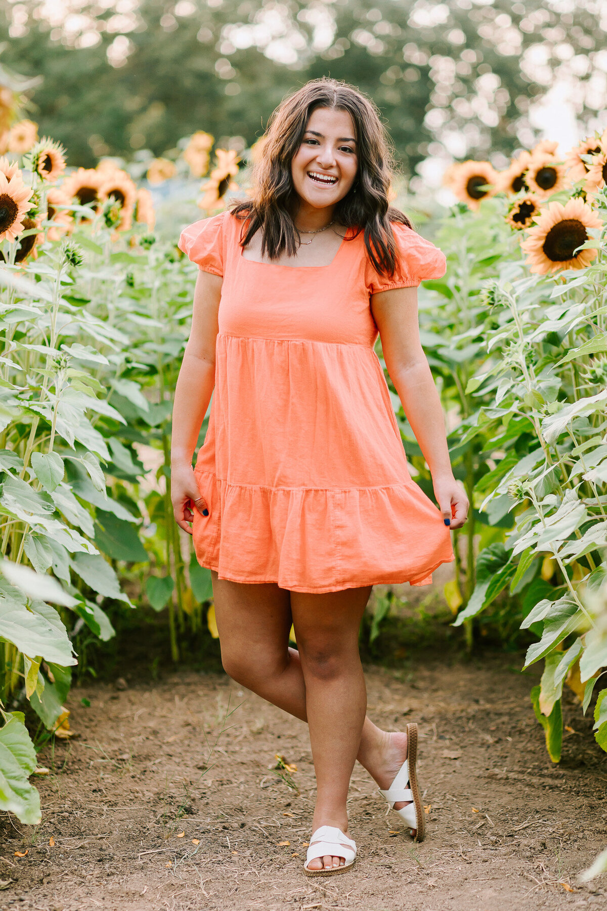 A teenage girl in a coral dress stands with one foot behind the other in a sunflower field