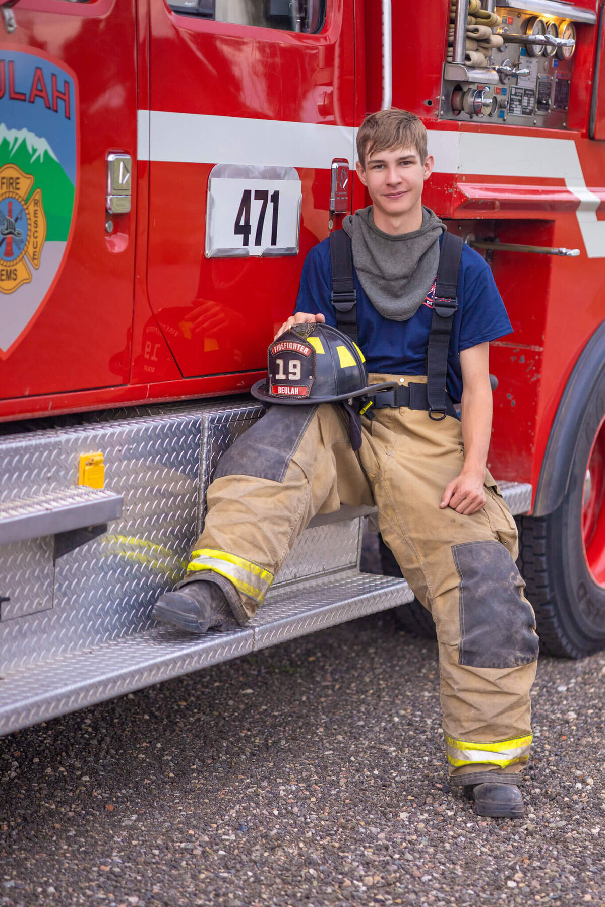 teenage boy in bunker gear holding his firefighter hat sitting on the side of a fire truck